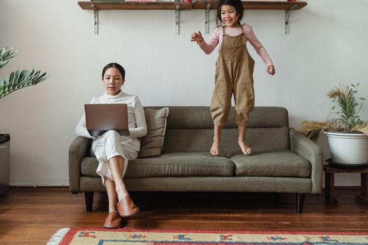 Image of a woman sitting on a couch looking at her laptop while her daughter jumps on the couch beside her