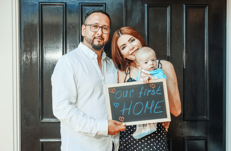 Image of a couple standing in front of a home holding a sign that says "Our First Home".