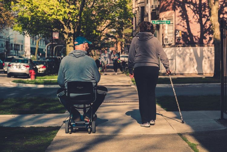 Image of a man sitting in a motorized scooter and a woman walking next to him holding a cane