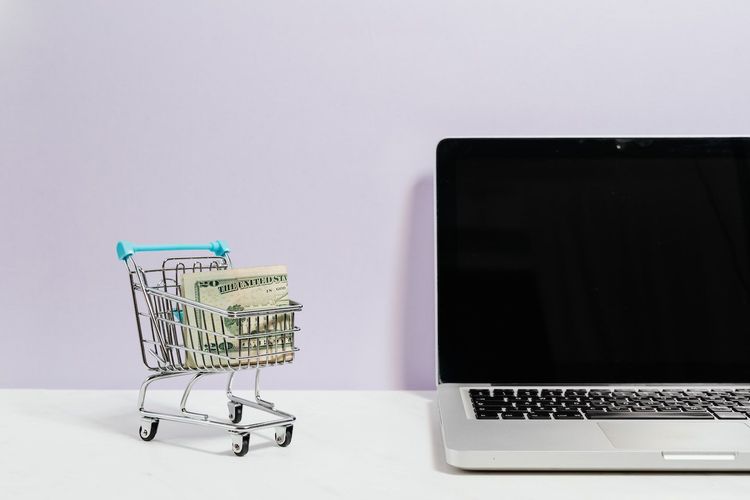 Image of a laptop on a desk and a miniature shopping cart with a twenty dollar bill beside it
