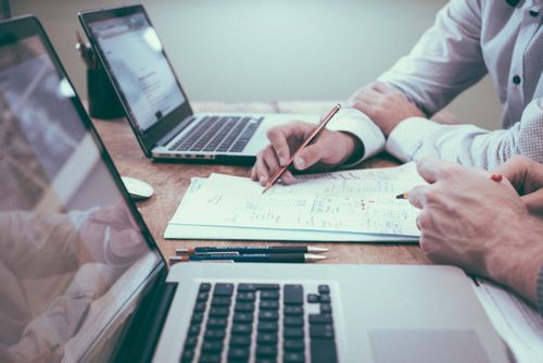 Image of two men looking at a piece of paper with two laptops in front of them