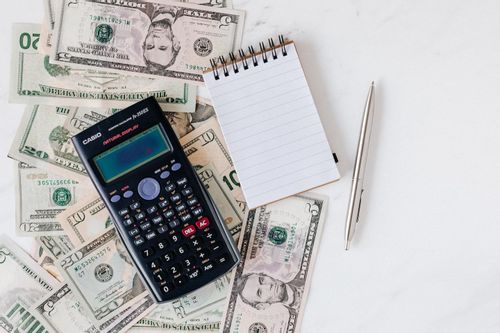 Image of a calculator, notepad, and pen sitting on a pile of five, ten, and twenty dollar bills