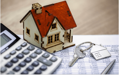 An image of a toy house, set of keys, piece of paper, calculator, and pen on top of a desk